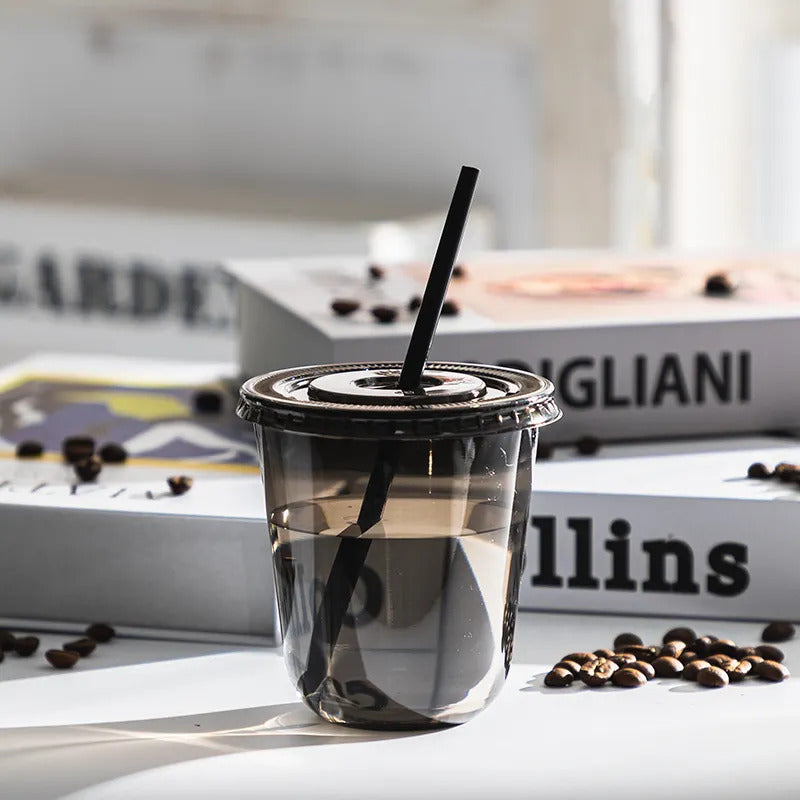 Clear plastic cup with a black straw on a surface with coffee beans and books in the background.